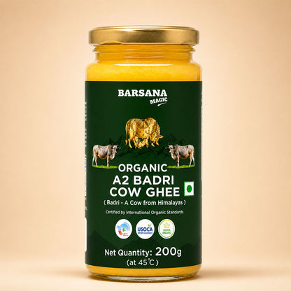 Barsana Magic and A 200g glass jar of Barsana Magic Organic A2 Badri Cow Ghee, filled with yellow ghee and featuring a dark green label with cow illustrations and a gold metal cap, set against a solid beige background.