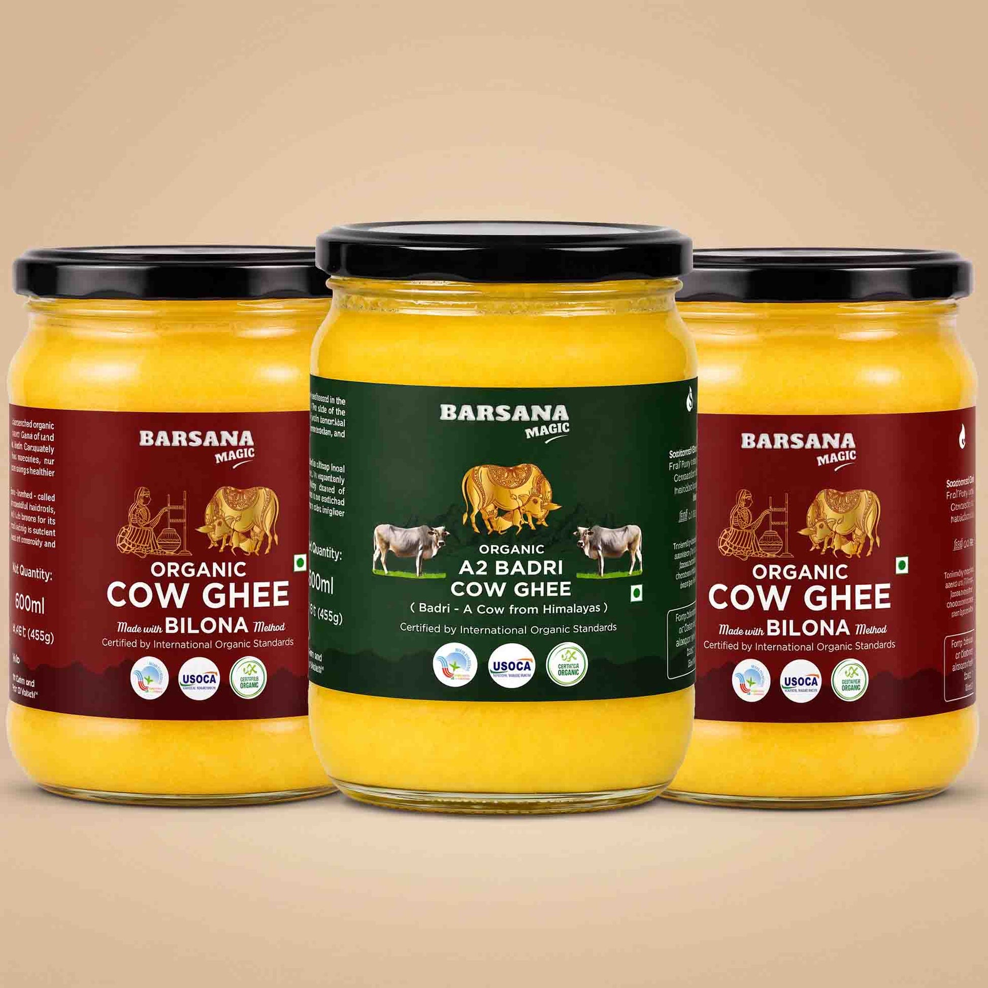 Barsana Magic and Three glass jars of Barsana Organic Cow Ghee arranged symmetrically on a warm beige background. The center jar has a dark green label, while the two side jars have dark red labels, all showing yellow ghee inside and black lids.