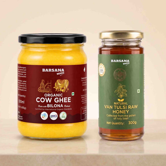 Barsana Magic and Two jars of Barsana products on a light beige surface against a warm beige background: a large jar of Organic Cow Ghee with a black lid and dark red label, and a smaller jar of Natural Van Tulsi Raw Honey with a gold lid and green label.