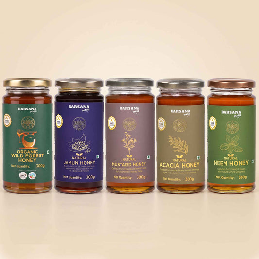 Barsana Magic and Five glass jars of Barsana Handmade honey arranged in a row on a light beige background. The jars have gold and silver metal lids and feature colorful labels in green, purple, brown, and olive tones, labeled with different honey varieties: Organic Wild Forest Honey, Natural Jamun Honey, Mustard Honey, Acacia Honey, and Neem Honey.