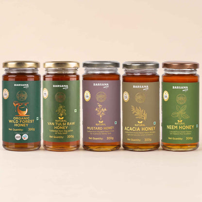 Barsana Magic and Five glass jars of brand honey arranged in a row against a light beige background. The jars contain amber-colored honey and have labels in green, purple, and brown tones with gold lettering.