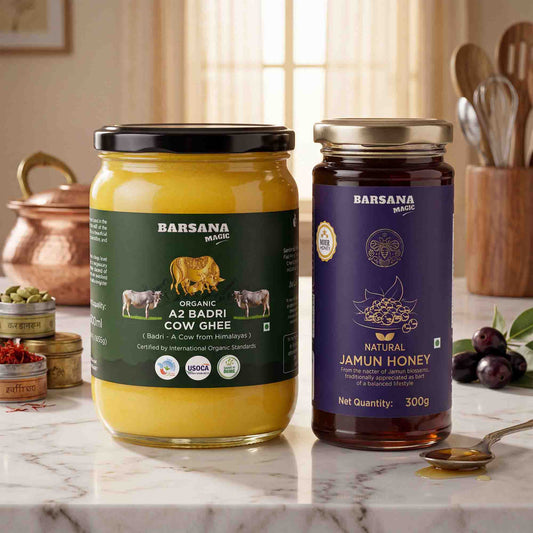 Barsana Magic and Two jars of Barsana Magic honey and ghee displayed on a marble countertop in a kitchen setting. The left jar contains yellow A2 Badri Cow Ghee with a black lid and green label, and the right jar contains natural jamun honey with a gold lid and purple label.
