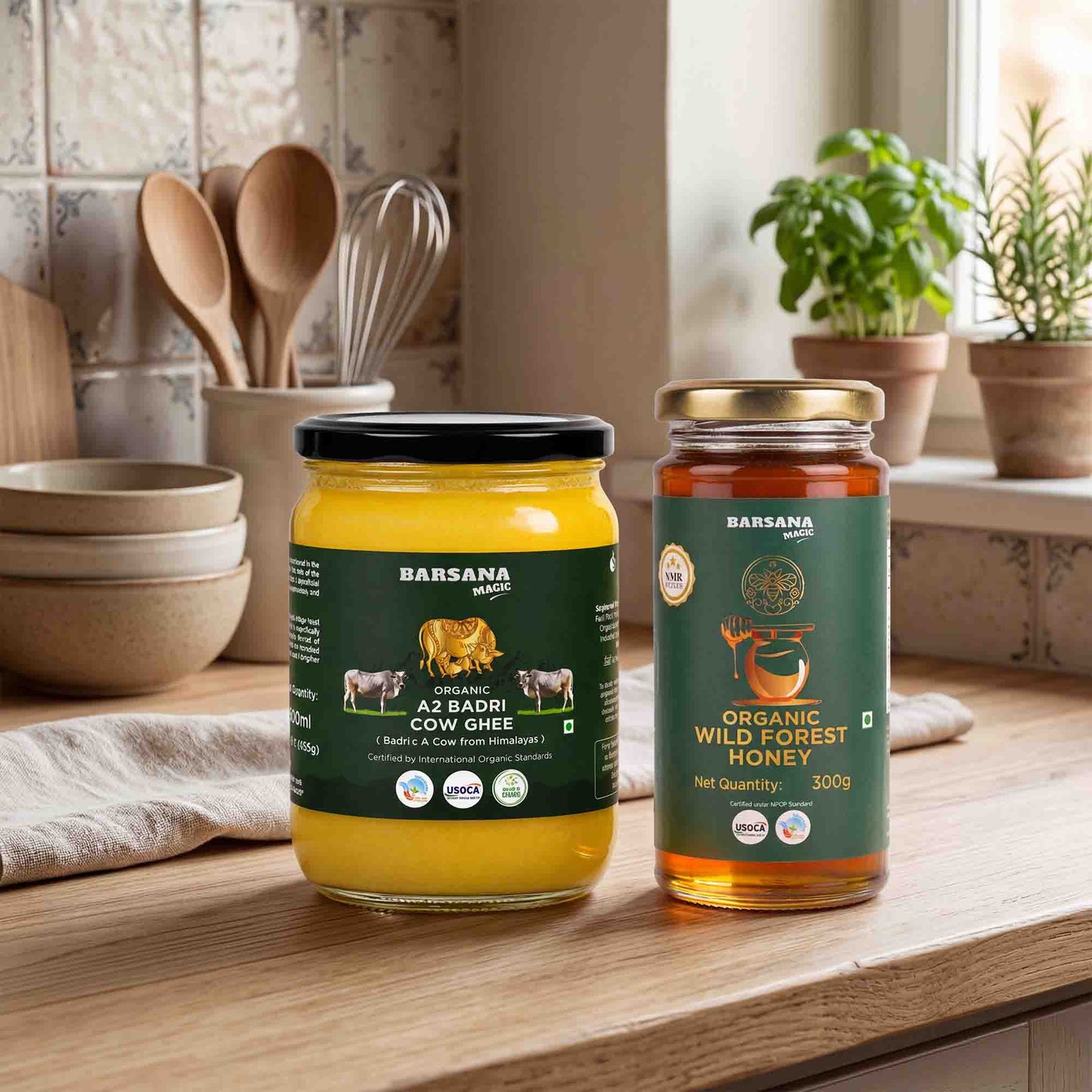 Barsana Magic and Two jars of Barsana ghee and honey displayed on a wooden kitchen counter. The left jar contains yellow A2 Badri Cow Ghee with a black lid and dark green label, and the right jar contains Organic Wild Forest Honey with a gold lid and dark green label.