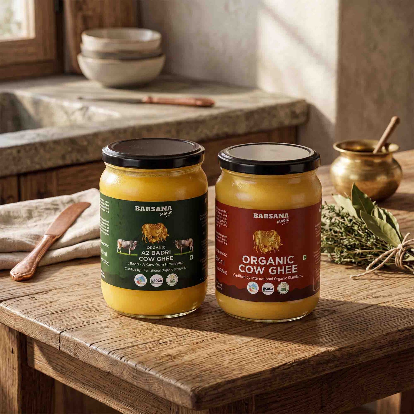 Barsana Magic and Two jars of Barsana organic cow ghee displayed on a rustic wooden table in a warm, traditional kitchen setting. One jar is labeled 'A2 Badhi Cow Ghee' with a dark green label, and the other is 'Organic Cow Ghee' with a dark red label.