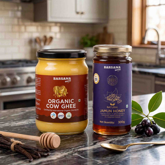 Barsana Magic and Two jars of Barsana brand honey and ghee displayed on a kitchen countertop with a wooden honey dipper, a spoon, and a small bunch of green leaves and dark berries. The left jar contains organic cow ghee with a black lid and a brown label, and the right jar contains natural jamun honey with a gold lid and a purple label.