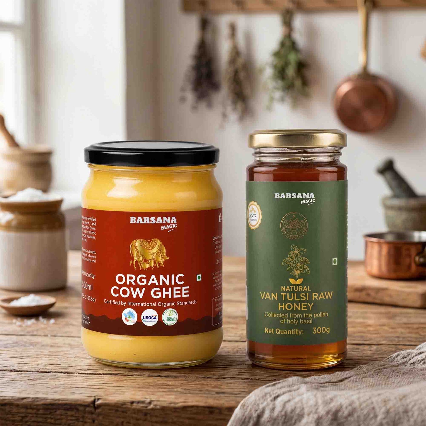 Barsana Magic and Two jars of Barsana Dairy products on a rustic wooden table: a jar of Organic Cow Ghee with a black lid and red label, and a jar of Natural Van Tulsi Raw Honey with a gold lid and green label. A kitchen scene with hanging copper pots is softly blurred in the background.