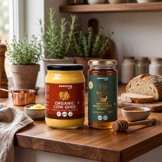 Barsana Magic and Two jars of Barsana ghee and honey displayed on a rustic wooden countertop in a warm kitchen setting, with bread, bowls, and potted plants in the background.