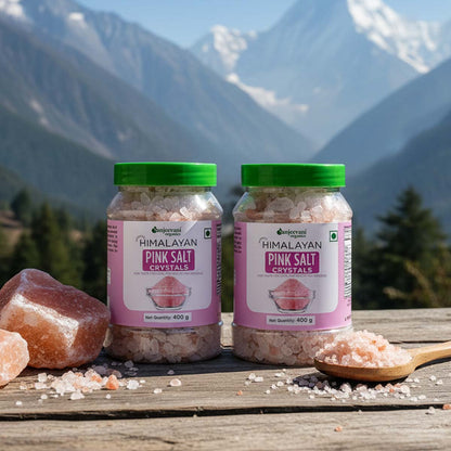 Barsana Magic and Two jars of Himalayan Pink Salt Crystals with pink salt crystals and a wooden spoon on a wooden surface with mountains in the background.