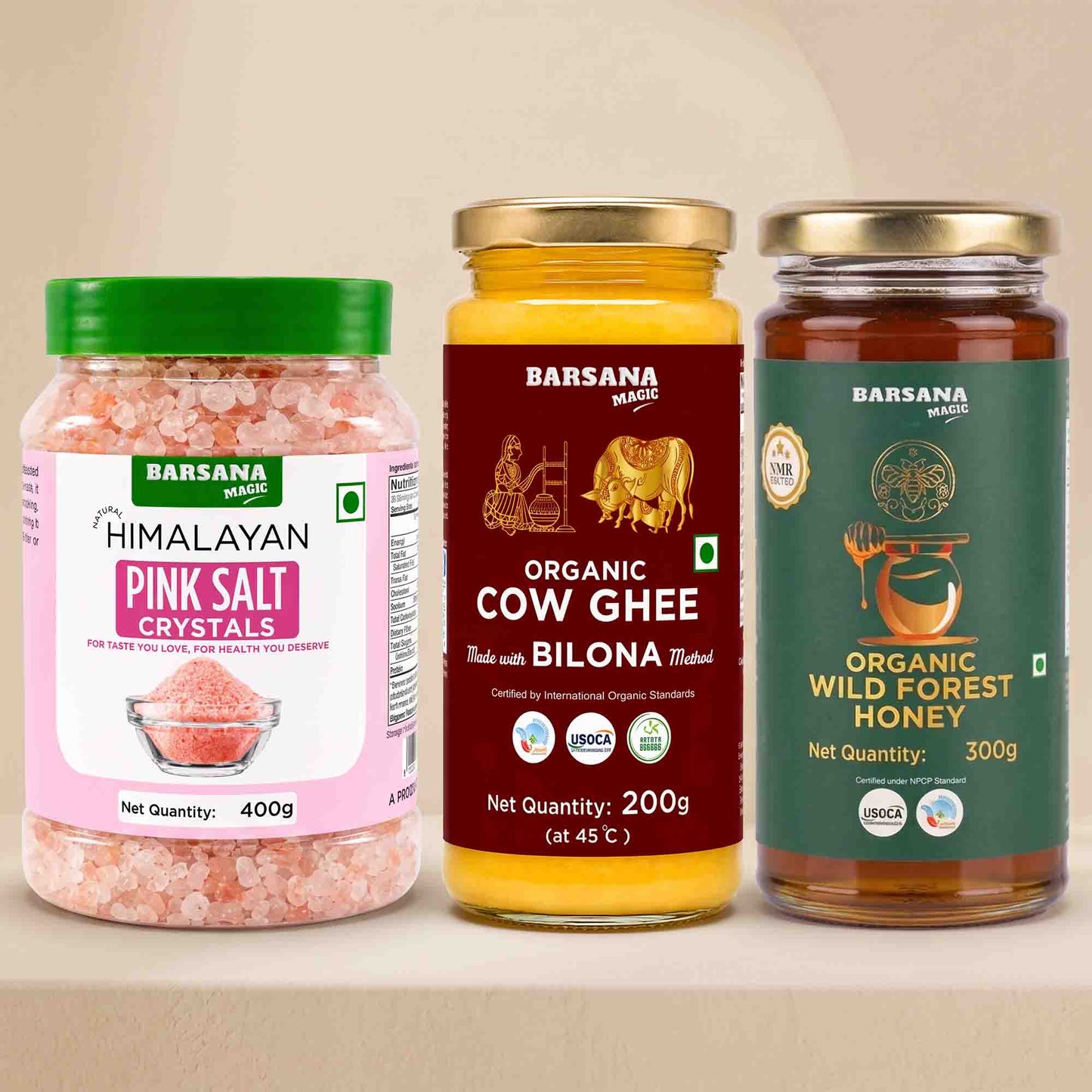 Barsana Magic and Three Barsana Magic products displayed side-by-side on a beige surface: Himalayan Pink Salt Crystals in a clear jar with a green lid, Organic Cow Ghee in a yellow jar with a gold lid, and Organic Wild Forest Honey in a dark green jar with a gold lid.