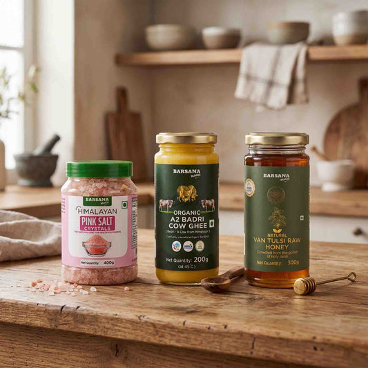 Barsana Magic and Three jars of Baransa brand food products arranged on a rustic wooden countertop in a warm, softly blurred kitchen setting: Himalayan Pink Salt Crystal, Organic A2 Badri Cow Ghee, and Van Tulsi Raw Honey. A wooden honey dipper and spoon are placed beside the jars.
