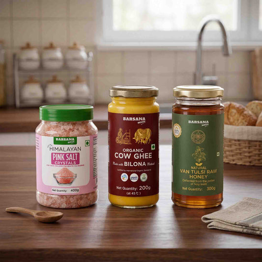 Barsana Magic and Three Barswana household products are displayed on a wooden countertop in a kitchen setting: Himalayan Pink Salt Crystals, Organic Cow Ghee, and Van Tulsi Raw Honey. The background shows a blurred kitchen scene with a sink and window.