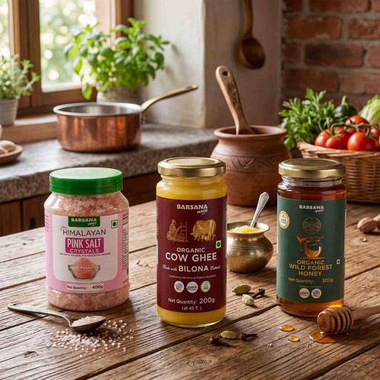 Barsana Magic and Three jars of Barsana brand food products are arranged on a rustic wooden table in a kitchen setting: Himalayan Pink Salt Geyser, Organic Cow Ghee, and Organic Wild Forest Honey. The scene includes cooking utensils, herbs, tomatoes, and a copper pot in the background.