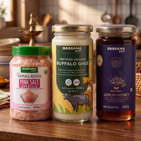 Barsana Magic and Three jars of Barsana Magic brand food products arranged on a wooden table: a pink salt crystal jar with a green lid, a buffalo ghee jar with a green and yellow label, and a jamun honey jar with a dark purple label.