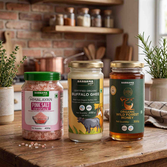 Barsana Magic and Three jars of Barsana brand food products arranged on a wooden table in a kitchen setting: Himalayan Pink Salt Crystals, Organic Buffalo Ghee, and Organic Wild Forest Honey. The background shows a brick wall, shelves with jars, and potted plants.